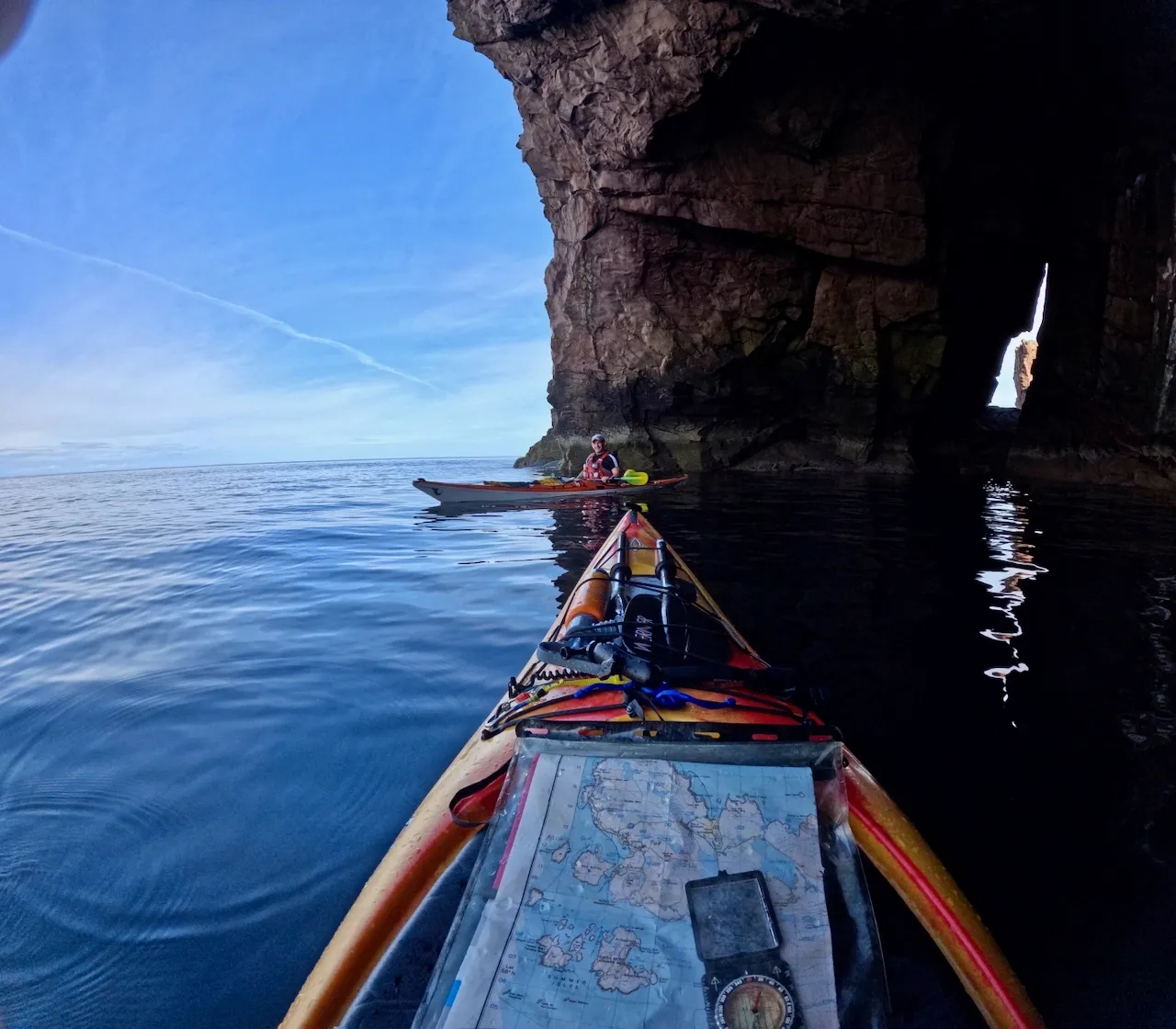 Sea kayak with map on splash deck, blue sea and sky, sea cave to the right second kayaker in the distance