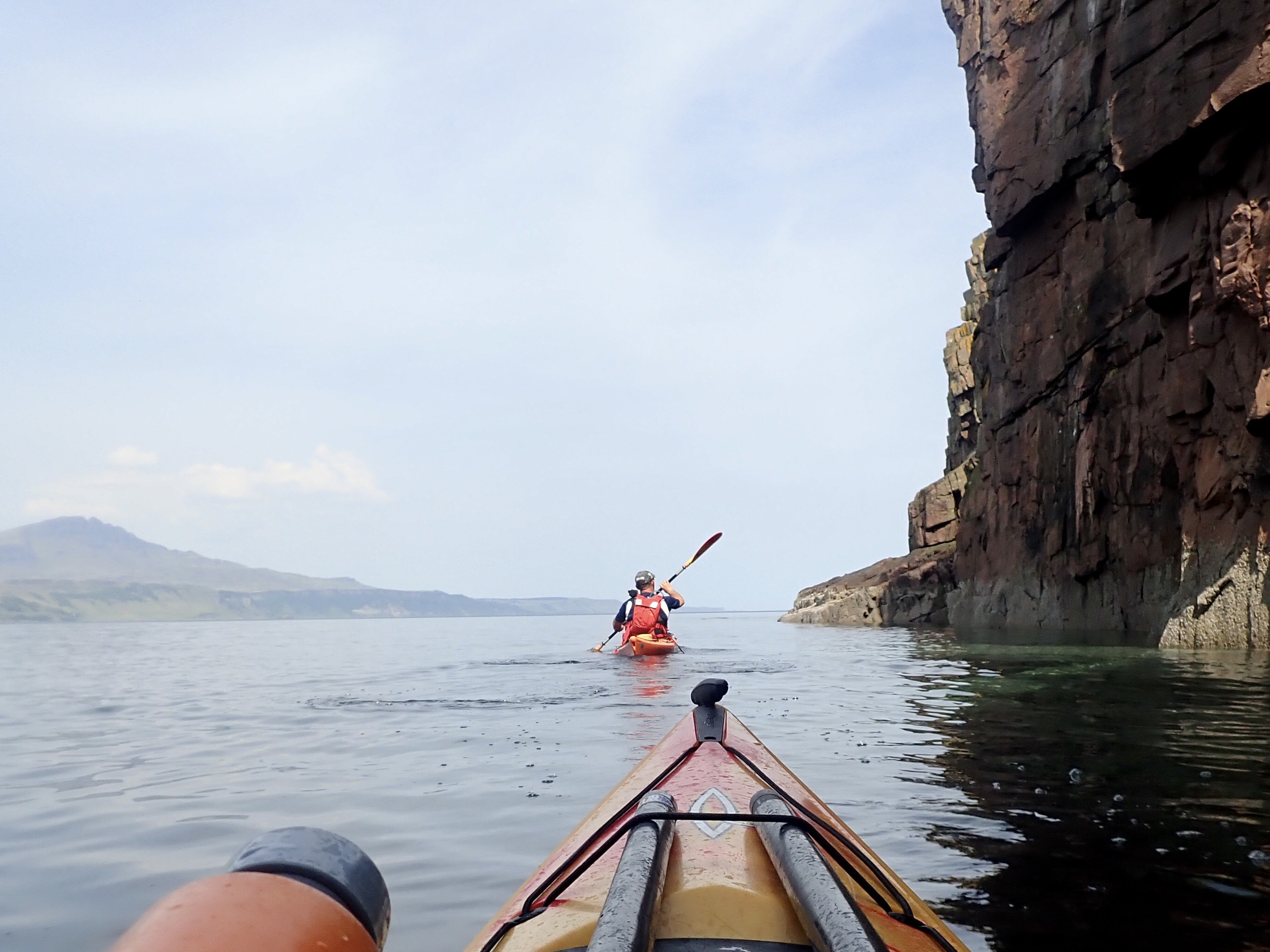 Two sea kayakers paddling beneath high coastal cliffs on calm water.